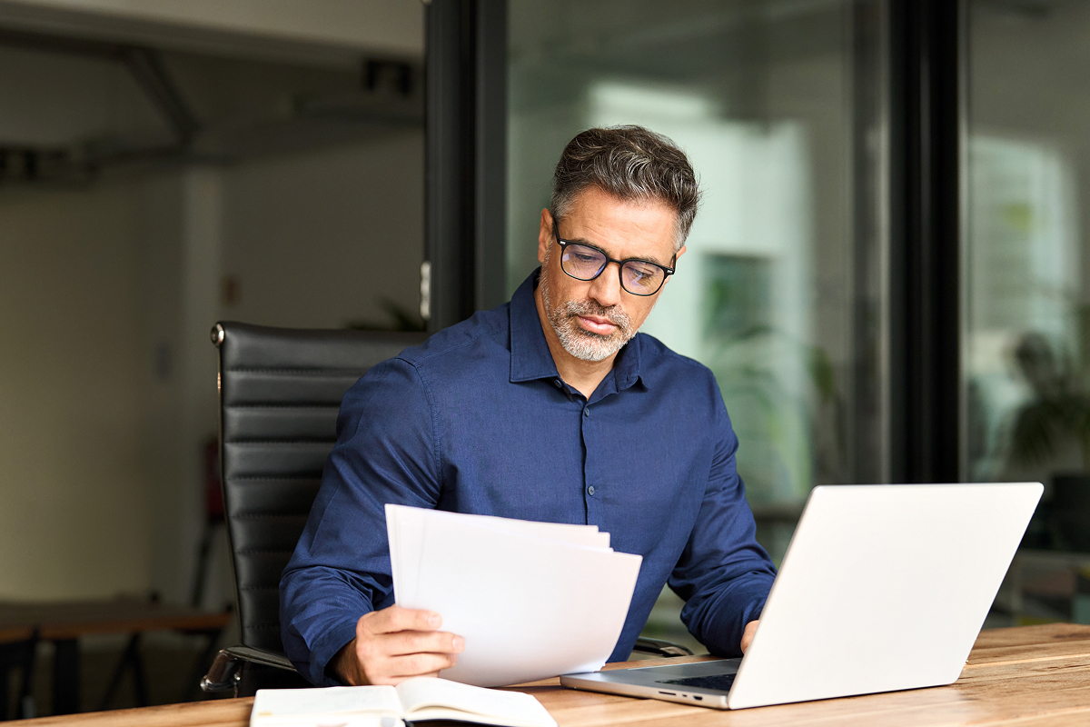 Man reading papers at a desk with an open laptop.