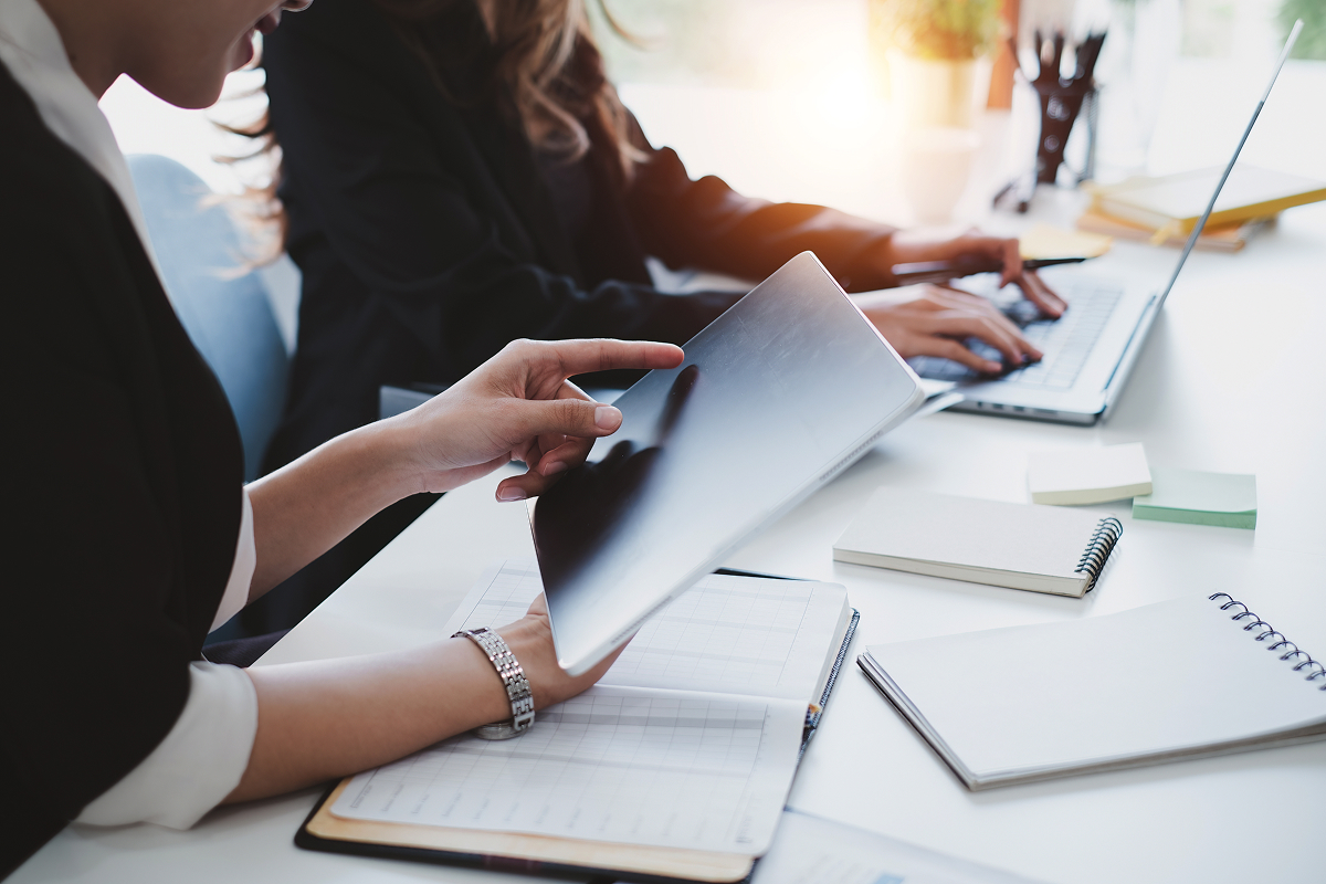 Two women working on a computer and tablet with notes on the table in front of them
